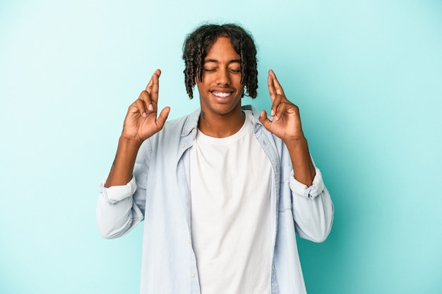 Jovem afro-americano isolado em um fundo azul cruzando os dedos para ter sorte