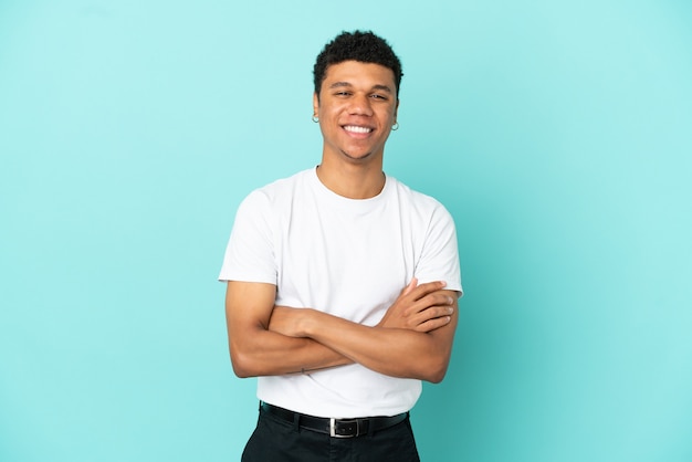 Jovem afro-americano isolado em um fundo azul, mantendo os braços cruzados na posição frontal