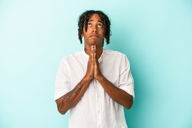 Jovem afro-americano isolado em um fundo azul, orando de mãos dadas perto da boca, sente-se confiante.