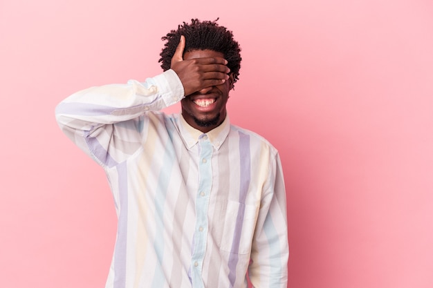 Jovem afro-americano isolado em um fundo rosa cobre os olhos com as mãos, sorri amplamente esperando por uma surpresa.