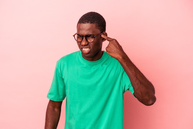 Jovem afro-americano isolado em um fundo rosa, cobrindo as orelhas com as mãos.