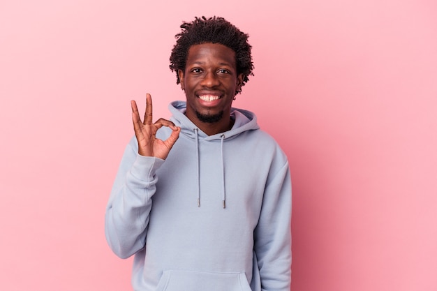 Jovem afro-americano isolado em um fundo rosa pisca um olho e segura um gesto de ok com a mão.