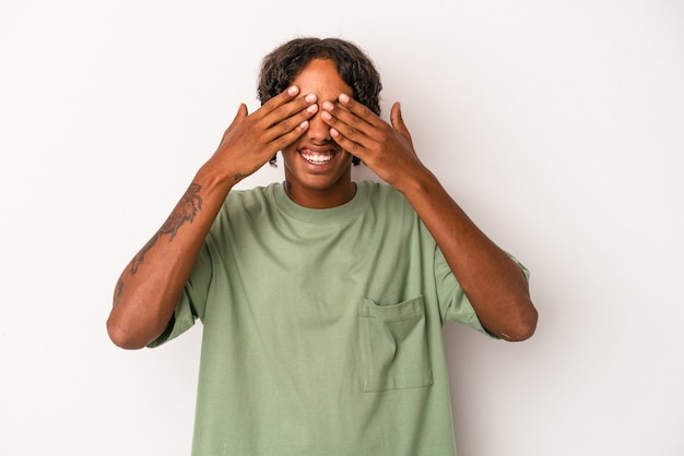 Jovem afro-americano isolado no fundo branco cobre os olhos com as mãos, sorri amplamente esperando por uma surpresa.