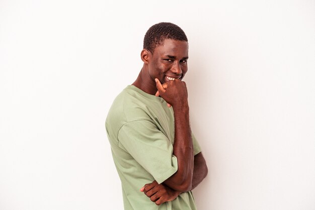 Jovem afro-americano isolado no fundo branco, sorrindo feliz e confiante, tocando o queixo com a mão.