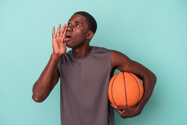 Jovem afro-americano jogando basquete isolado em fundo azul gritando e segurando a palma da mão perto da boca aberta