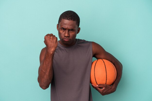 Jovem afro-americano jogando basquete isolado em fundo azul, mostrando o punho para a expressão facial agressiva da câmera