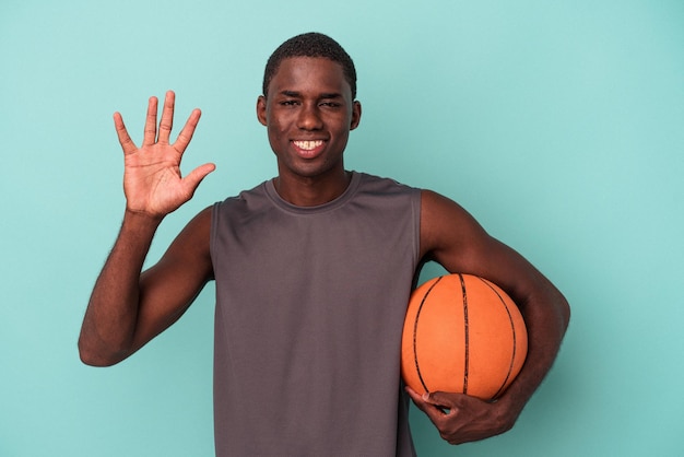 Jovem afro-americano jogando basquete isolado em fundo azul sorrindo alegre mostrando o número cinco com os dedos
