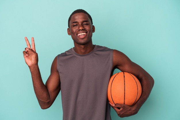 Jovem afro-americano jogando basquete isolado em um fundo azul, alegre e despreocupado, mostrando um símbolo de paz com os dedos.