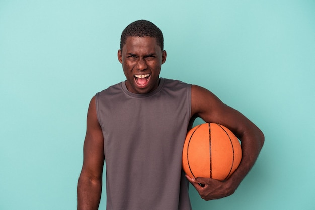 Jovem afro-americano jogando basquete isolado no fundo azul, gritando muito irritado e agressivo.