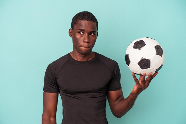 Jovem afro-americano jogando futebol isolado em fundo azul confuso, sente-se duvidoso e inseguro.