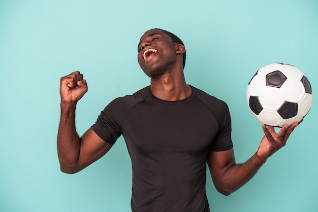 Jovem afro-americano jogando futebol isolado em fundo azul, levantando o punho após uma vitória, conceito de vencedor.