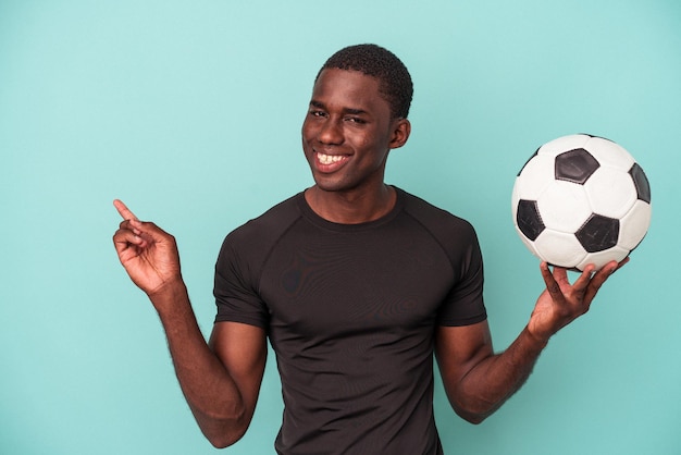 Jovem afro-americano jogando futebol isolado em fundo azul, sorrindo e apontando de lado, mostrando algo no espaço em branco.