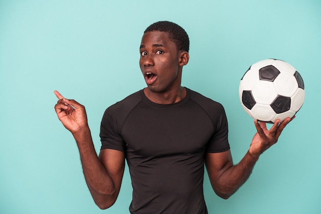 Jovem afro-americano jogando futebol isolado em um fundo azul apontando para o lado