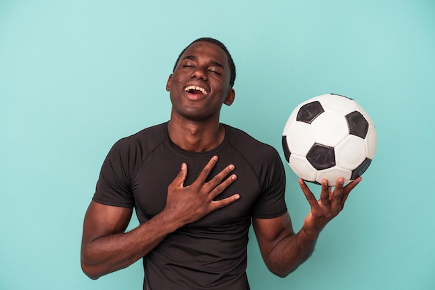 Jovem afro-americano jogando futebol isolado no fundo azul ri alto, mantendo a mão no peito.