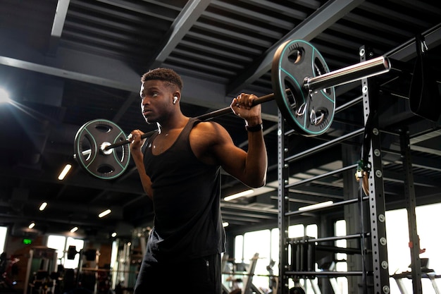 Jovem afro-americano levanta barra no ginásio escuro, cara de esportes treina na sala de ginástica