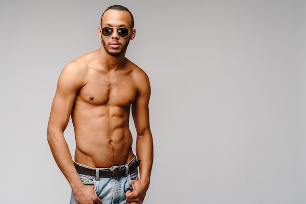 Foto jovem afro-americano musculoso sem camisa usando óculos escuros sobre a parede cinza claro