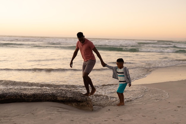 Jovem afro-americano segurando a mão do filho enquanto caminhava na praia contra o céu claro ao pôr do sol