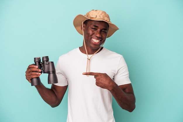 Jovem afro-americano segurando binóculos isolados em um fundo azul, sorrindo e apontando de lado, mostrando algo no espaço em branco.