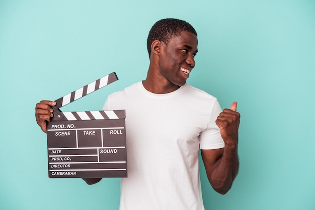 Jovem afro-americano segurando claquete isolada em fundo azul aponta com o dedo polegar longe, rindo e despreocupado.