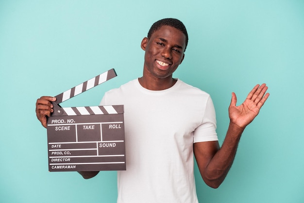 Jovem afro-americano segurando claquete isolada em fundo azul, mostrando um espaço de cópia na palma da mão e segurando a outra mão na cintura.