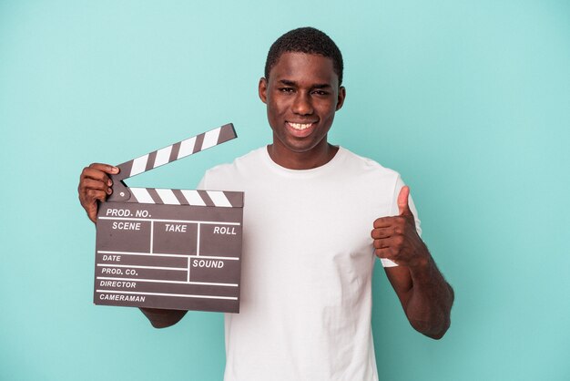 Jovem afro-americano segurando claquete isolada em um fundo azul, sorrindo e levantando o polegar