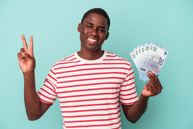 Jovem afro-americano segurando notas de banco isoladas em fundo azul alegre e despreocupada, mostrando um símbolo de paz com os dedos