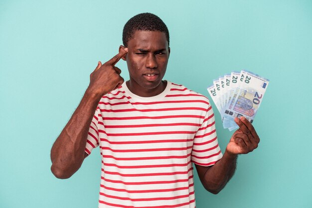 Jovem afro-americano segurando notas de banco isoladas em fundo azul, mostrando um gesto de decepção com o dedo indicador.