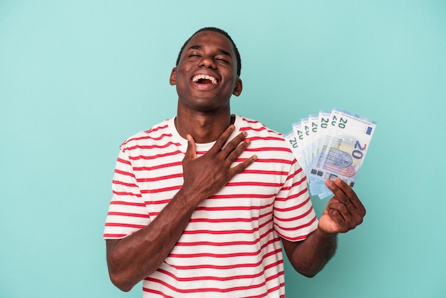 Jovem afro-americano segurando notas de banco isoladas em fundo azul ri alto, mantendo a mão no peito.