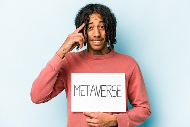 Jovem afro-americano segurando o cartaz do metaverso isolado em fundo azul, mostrando um gesto de decepção com o dedo indicador