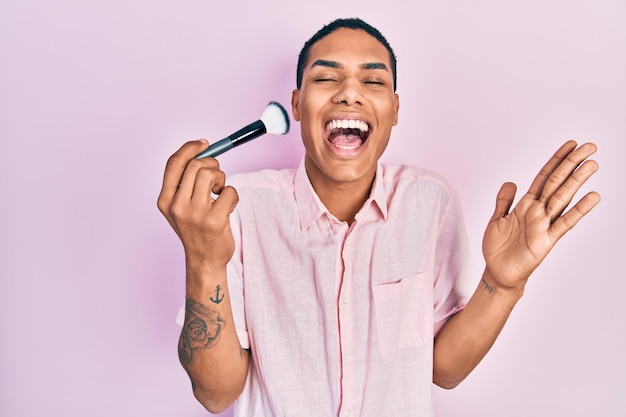 Jovem afro-americano segurando o pincel de maquiagem comemorando a conquista com sorriso feliz e expressão vencedora com a mão erguida