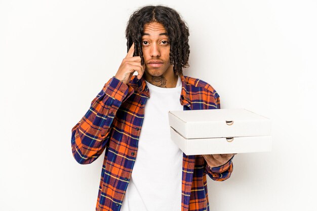 Jovem afro-americano segurando pizzas isoladas em fundo azul apontando o templo com o dedo pensando focado em uma tarefa