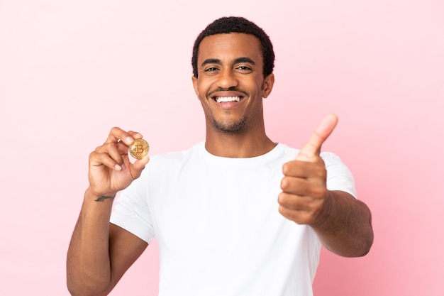 Jovem afro-americano segurando um bitcoin sobre um fundo rosa isolado com o polegar para cima porque algo bom aconteceu