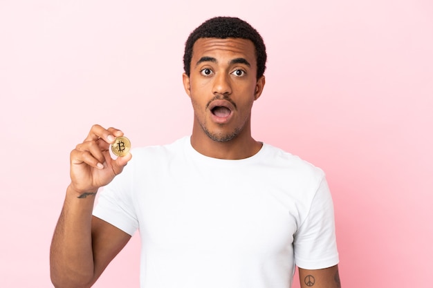 Jovem afro-americano segurando um Bitcoin sobre um fundo rosa isolado com uma expressão facial surpresa