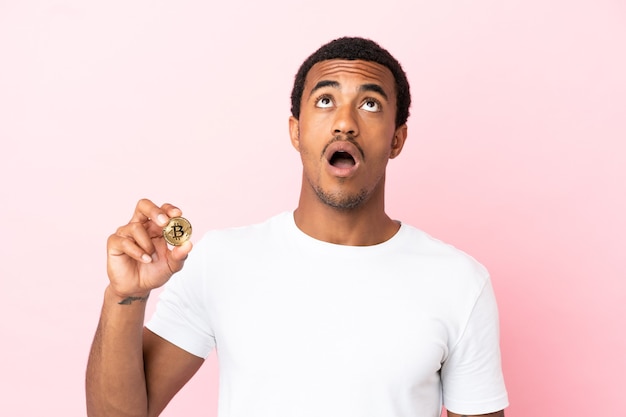 Jovem afro-americano segurando um Bitcoin sobre um fundo rosa isolado, olhando para cima e com expressão de surpresa