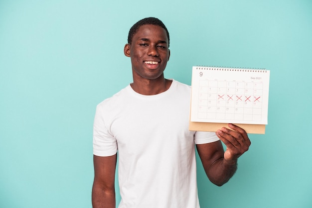 Jovem afro-americano segurando um calendário isolado em fundo azul feliz, sorridente e alegre.