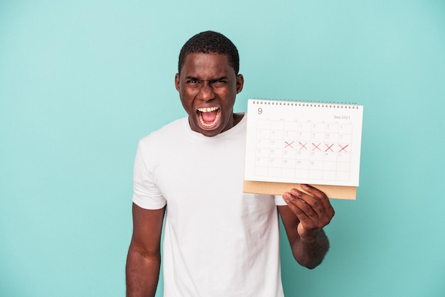 Jovem afro-americano segurando um calendário isolado em fundo azul, gritando muito irritado e agressivo.