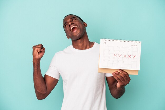 Jovem afro-americano segurando um calendário isolado no fundo azul, levantando o punho após uma vitória, o conceito de vencedor.