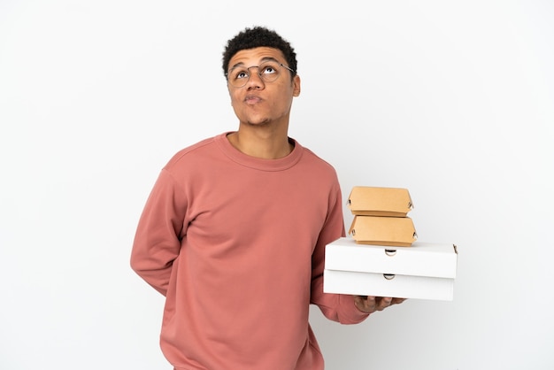 Jovem afro-americano segurando um hambúrguer e pizzas isoladas no fundo branco e olhando para cima