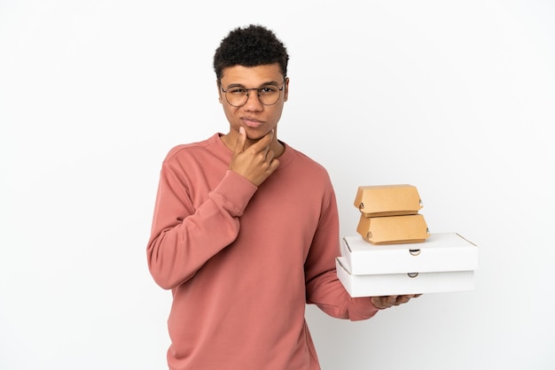 Jovem afro-americano segurando um hambúrguer e pizzas isoladas no fundo branco pensando