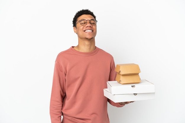 Jovem afro-americano segurando um hambúrguer e pizzas isoladas no fundo branco rindo