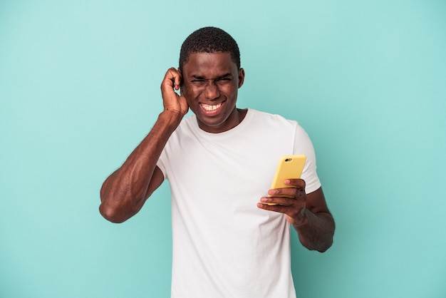 Jovem afro-americano segurando um telefone celular isolado em um fundo azul, cobrindo as orelhas com as mãos.