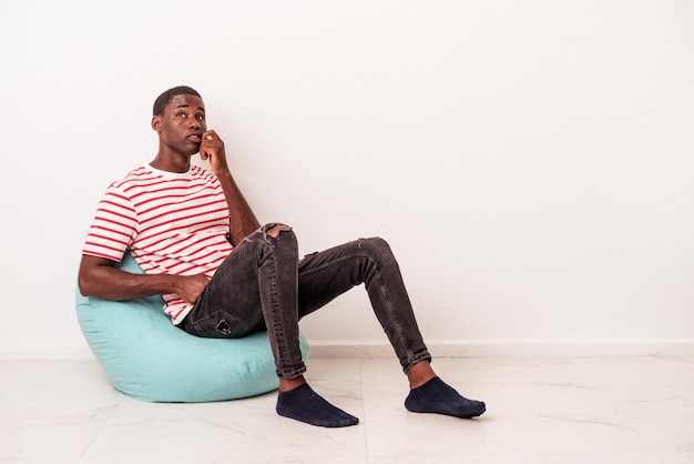 Jovem afro-americano sentado em um sopro isolado no fundo branco mordendo as unhas nervoso e muito ansioso