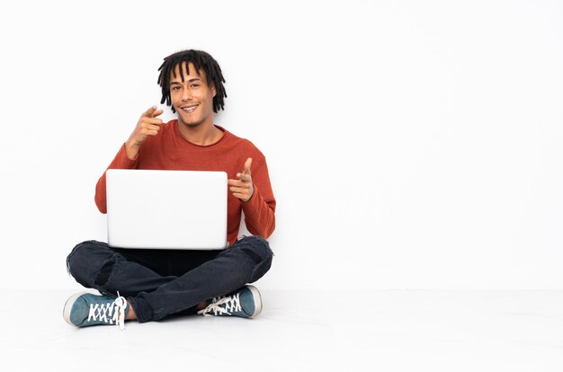 Foto jovem afro-americano sentado no chão e trabalhando com o laptop apontando para a frente e sorrindo