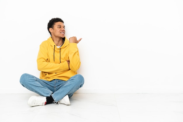 Jovem afro-americano sentado no chão, isolado no fundo branco, apontando para o lado para apresentar um produto