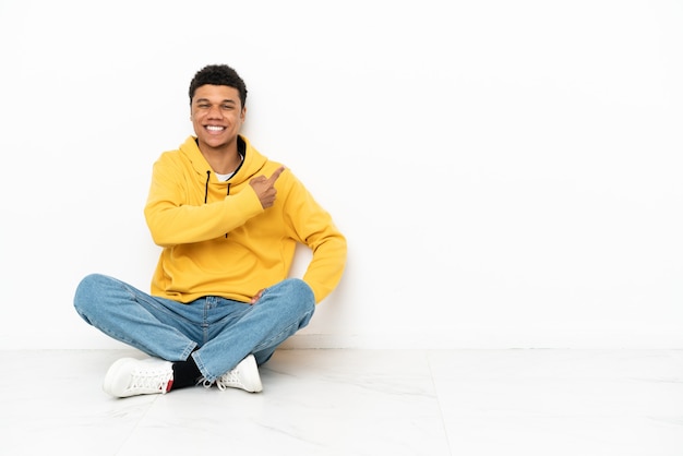 Jovem afro-americano sentado no chão, isolado no fundo branco, apontando para o lado para apresentar um produto