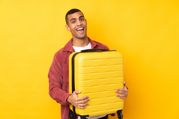 Jovem afro-americano sobre parede amarela isolada em férias com mala de viagem e surpreso