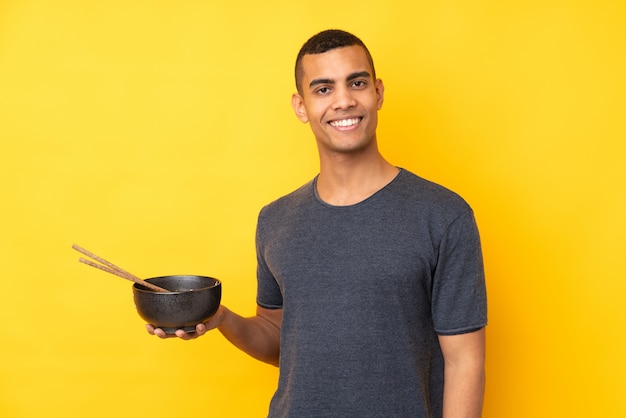 Jovem afro-americano sobre parede amarela isolada, sorrindo muito, segurando uma tigela de macarrão com pauzinhos