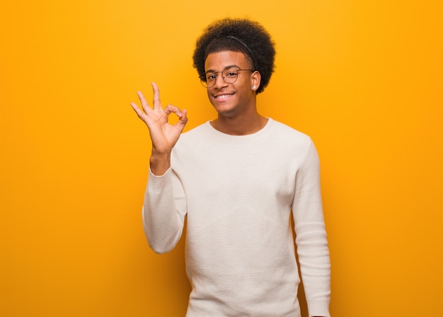 Jovem afro-americano sobre uma parede laranja alegre e confiante, fazendo o gesto de ok