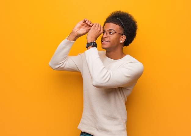 Jovem afro-americano sobre uma parede laranja, fazendo o gesto de uma luneta