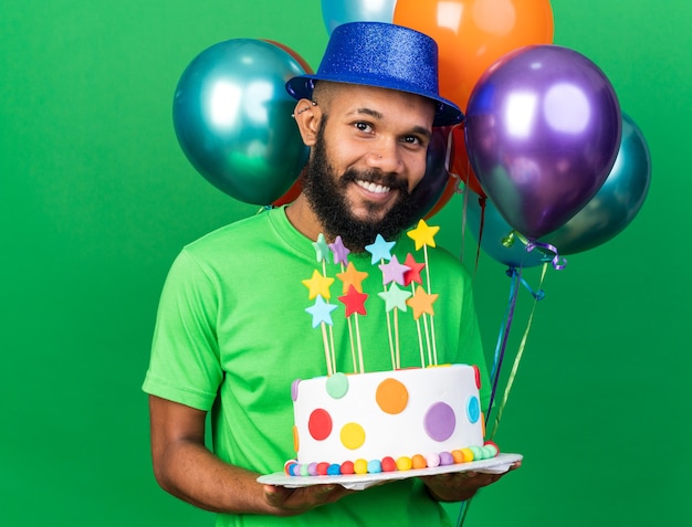 Foto jovem afro-americano sorridente com chapéu de festa parado na frente de balões segurando um bolo